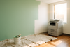 create image of an office, with a wall being painted. 3/4 of the wall is painted pastel green with the other 1/4 white. The canvas drop cloth is covering the hardwood floors. Only a small section of floor is not covered, near a  large printer with a window above it to the right. A lens flare shining through the window. A paint bucket on the canvas with a paint brush lying on it. 